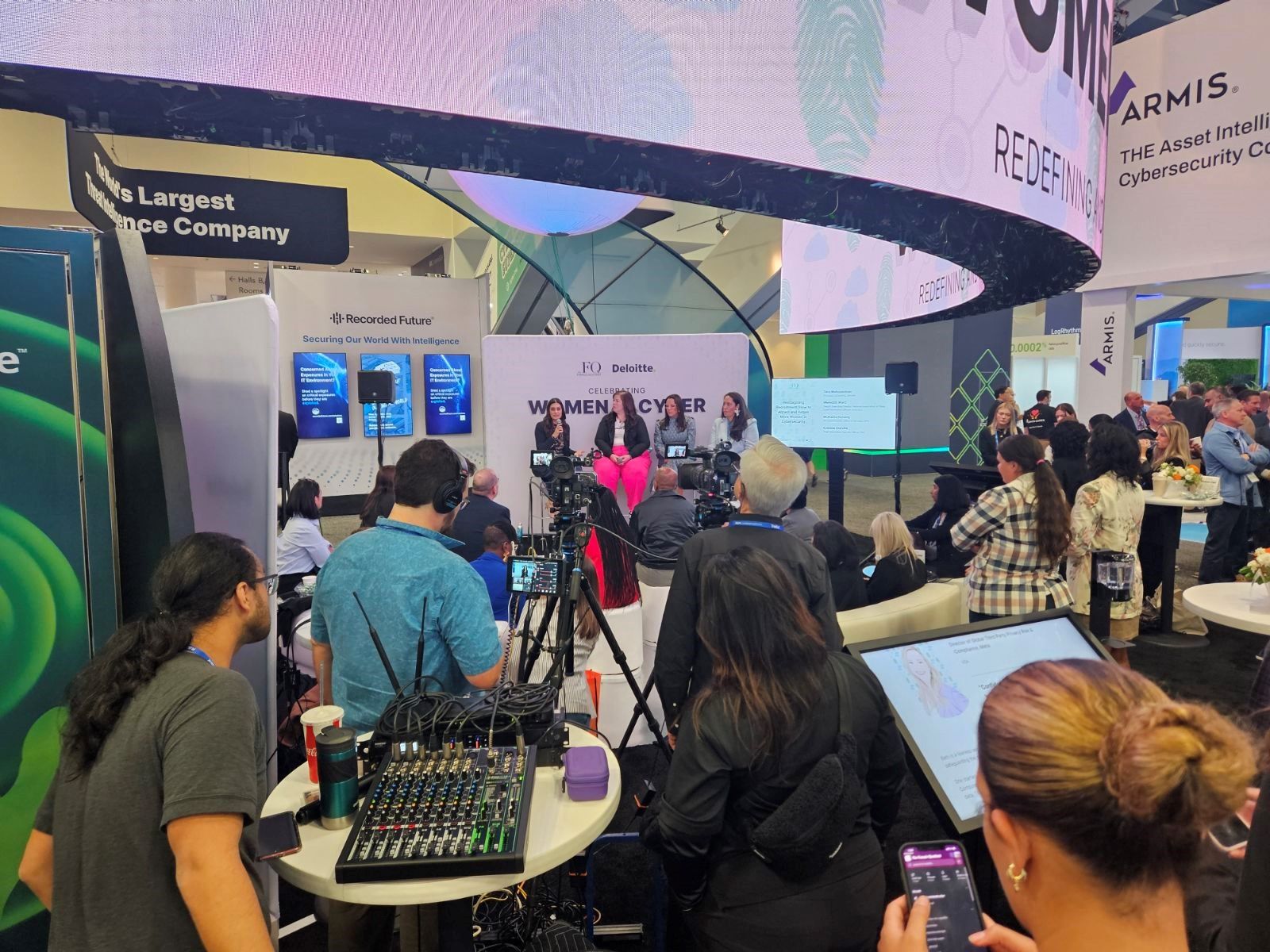 People attend a "Women in Cyber" panel talk at a conference. The room is filled with cameras, tech equipment, and attendees