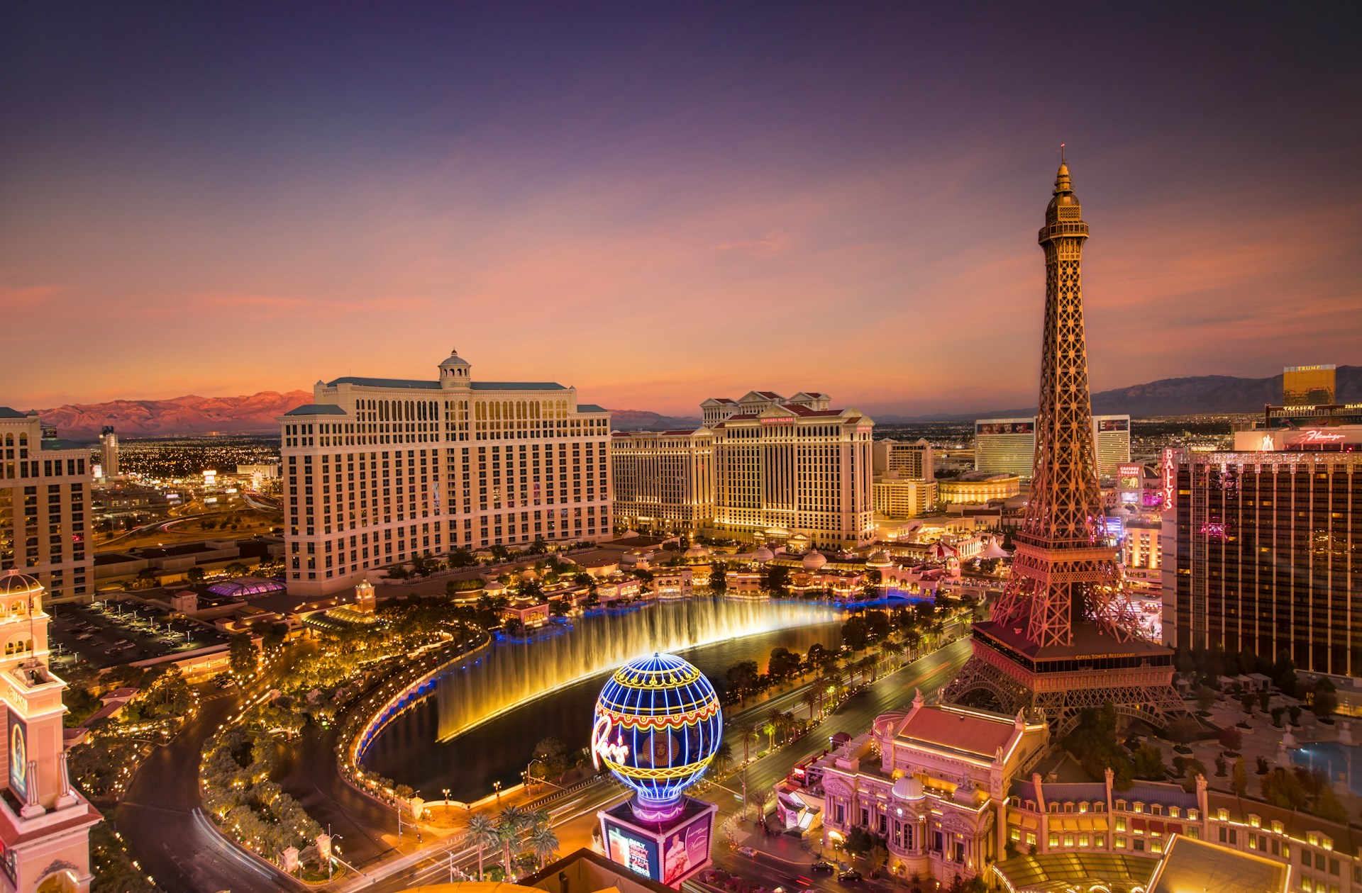 Aerial view of Las Vegas at sunset, featuring the illuminated Eiffel Tower replica, Bellagio Hotel, and fountain, with a vibrant, colorful skyline.