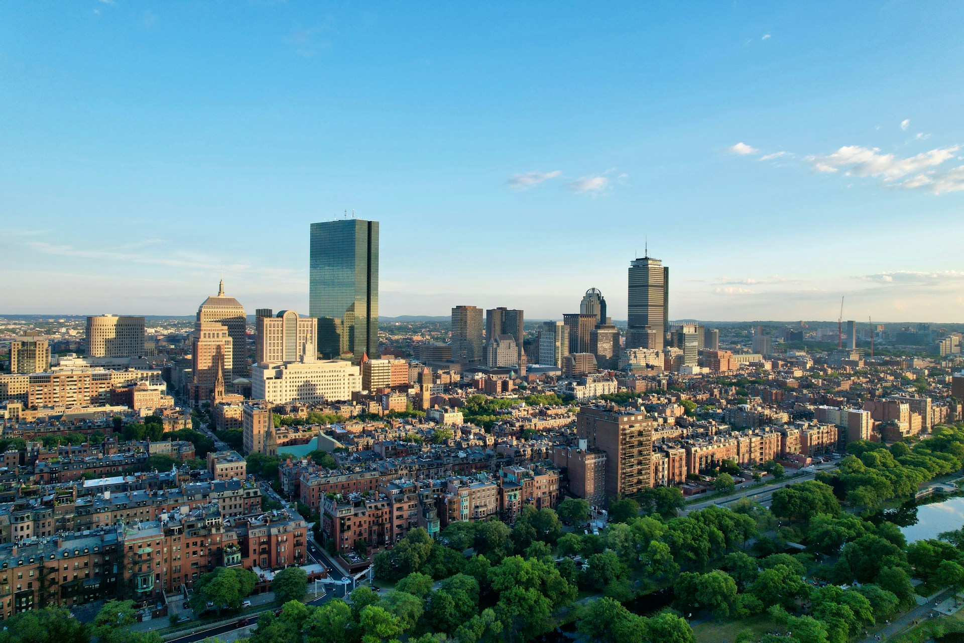 Aerial view of a city skyline at sunset with modern skyscrapers and historic buildings, surrounded by lush greenery and a clear blue sky.