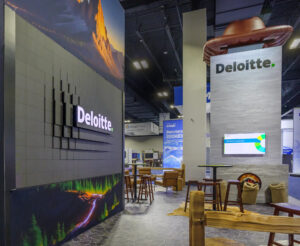 Exhibition booth featuring Deloitte branding, with large digital screens and seating area