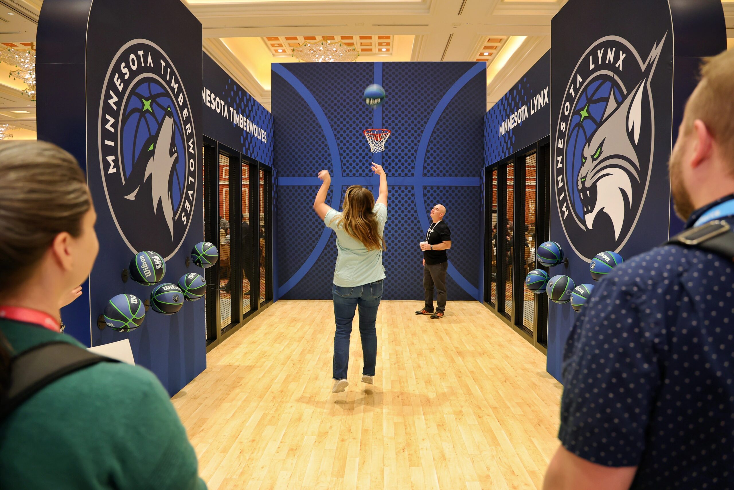A woman shoots a basketball in a sports-themed area with Minnesota Timberwolves and Lynx logos.