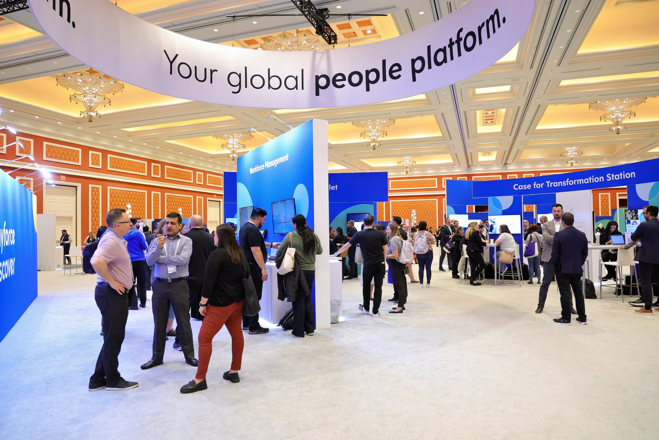 A busy conference hall with people networking and interacting at various booths. A large overhead banner reads "Your global people platform."